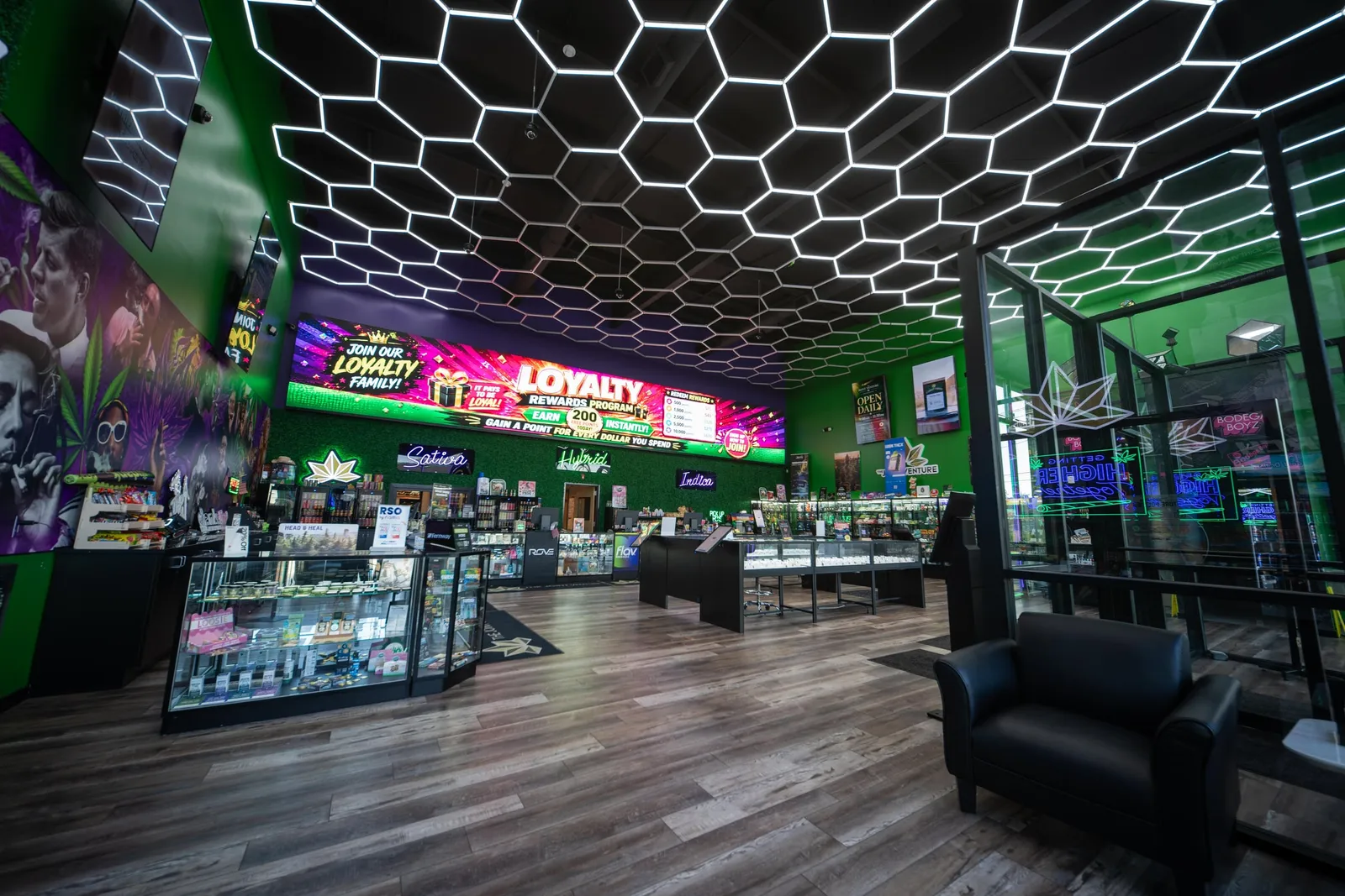 The showroom under the hex-grid LED ceiling, with the Loyalty screen and the Sativa, Hybrid and Indica neons along the back wall.